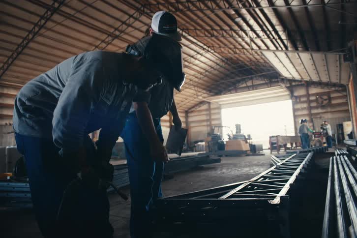 A supervisor inspects a metal truss and gives instructions to a welder in a large fabrication workshop