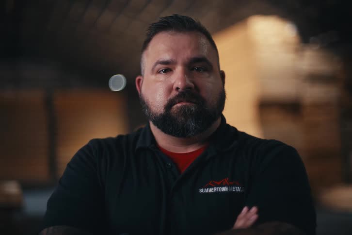 A confident and serious bearded man with arms crossed looks directly at the camera inside an industrial warehouse