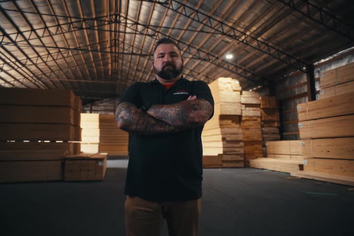 Portrait of a serious man with a beard and tattoos standing with arms crossed in a large lumber warehouse