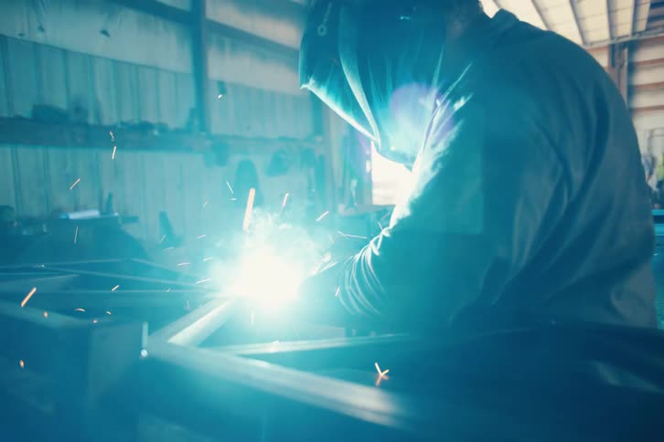 Dynamic tracking shot of a welder in a protective helmet working on a steel frame as bright sparks fly in a bustling industrial workshop