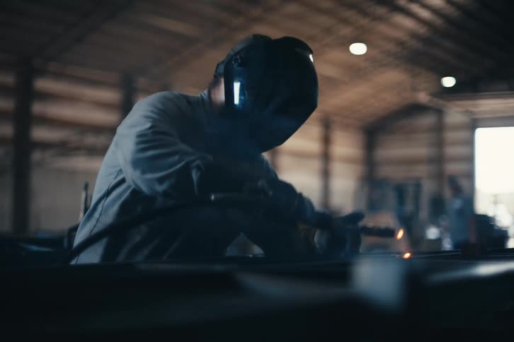 A skilled metalworker in a protective mask uses a welding torch, creating bright sparks in a dimly lit industrial workshop