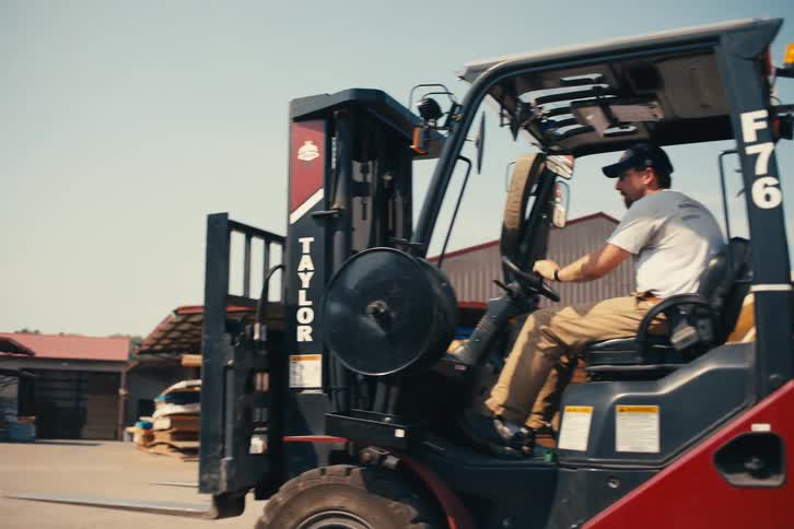 A forklift operator drives away from a truck loaded with lumber in a supply yard