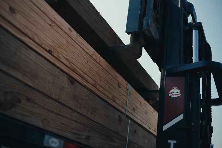 A close-up, low-angle shot shows a forklift slowly lowering a stack of wooden planks onto a flatbed trailer