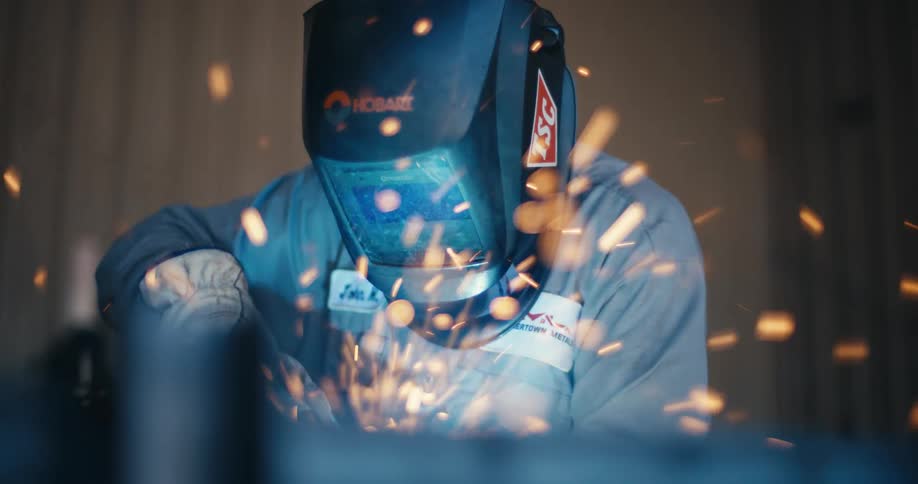A skilled metalworker welds a piece of steel in slow motion, creating a shower of bright sparks