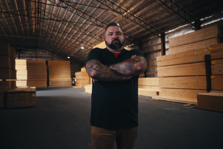 A confident man with tattoos and a beard crosses his arms and stares at the camera inside a large lumber warehouse