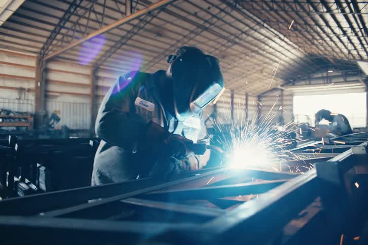 A skilled metalworker in a large industrial factory welds a steel frame, with bright blue light and sparks flying from the torch