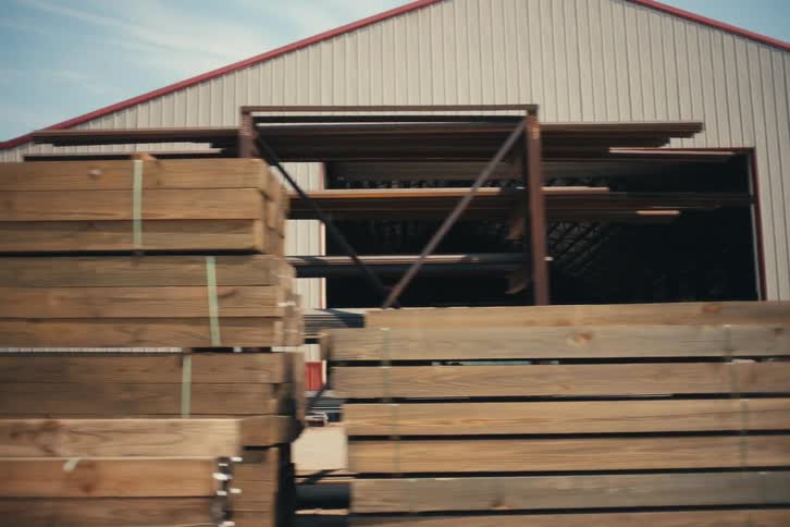 Slow pan past stacks of treated lumber and steel beams stored outside an industrial warehouse on a sunny day