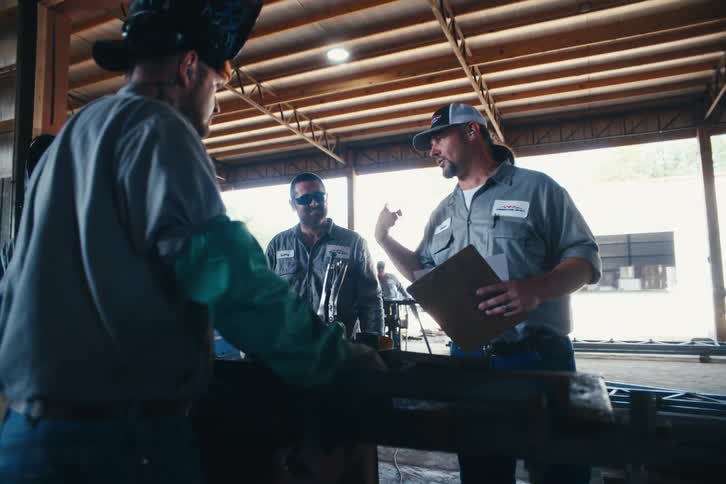 A supervisor with a clipboard gives instructions to his team of welders in a large metal fabrication workshop