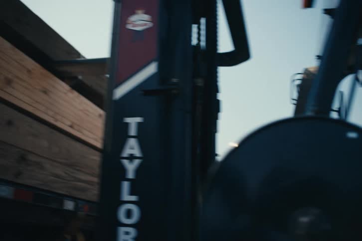 A forklift operator in slow motion carefully drives alongside a flatbed truck loaded with construction lumber