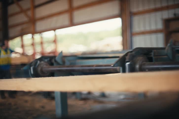 Two lumber mill workers in safety vests lift a freshly cut wooden plank onto a conveyor belt in slow motion