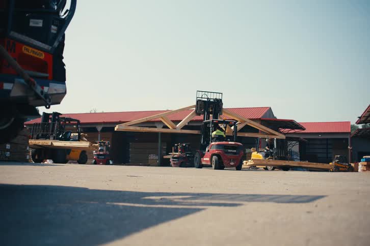 A red forklift carefully lifts and moves a large wooden roof truss at an industrial lumber yard on a sunny day