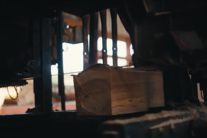 A wideband industrial sawmill slowly slices through a large block of wood, creating sawdust and lumber