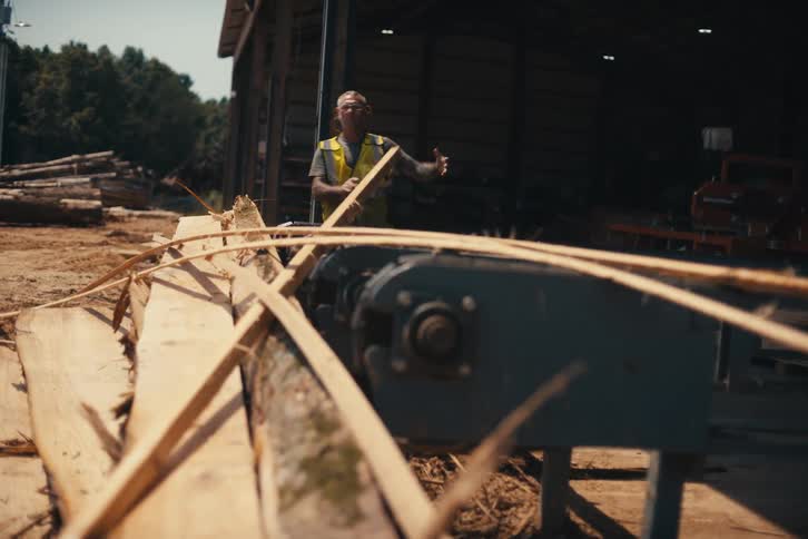 A hardworking man in a safety vest clears wood scraps from a conveyor belt at a rural lumber mill