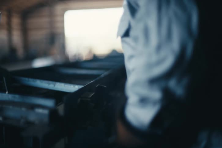 A metalworker in a welding helmet clamps a steel bar into place in a slow-motion shot at an industrial fabrication shop