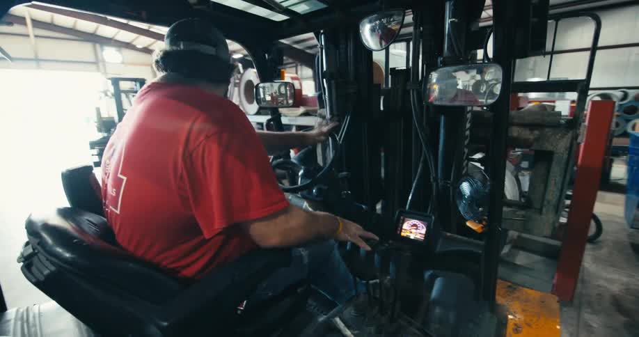 A forklift operator with a beard drives his vehicle through a warehouse while looking at the camera in a selfie-style shot