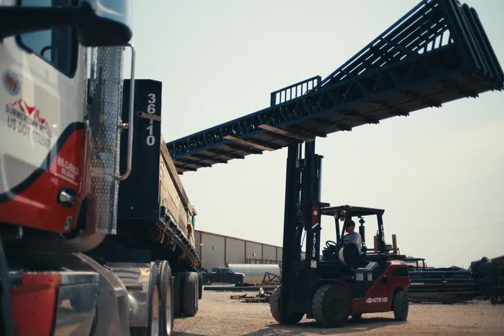 A forklift operator lifts a long metal frame high into the air next to a semi-truck at a construction supply yard