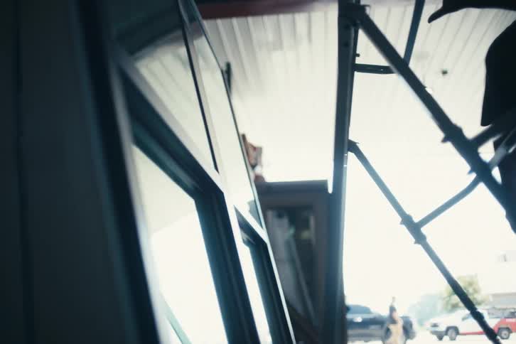 A silhouetted worker moves a large window on a dolly across a concrete floor in a sunlit industrial warehouse