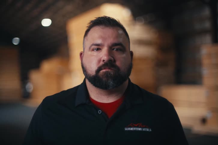 A serious bearded man stands proudly in a large warehouse filled with stacked lumber
