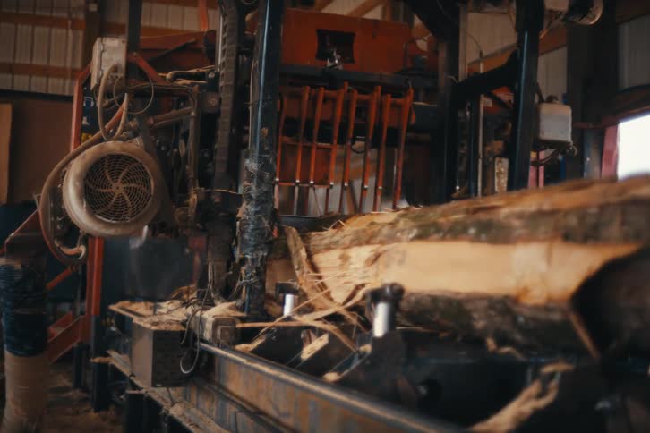 A slow-motion push-in shot follows a large log as it is being cut by an industrial sawmill in a workshop