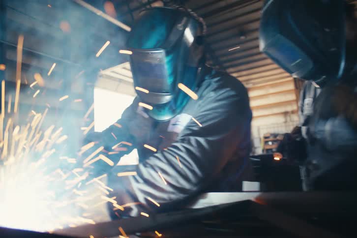 Close up slow motion dolly shot of two welders working side-by-side in a metal shop, creating a shower of bright sparks
