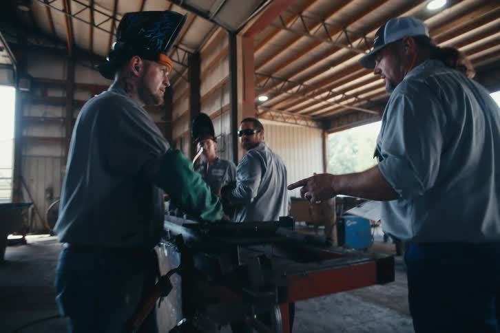 A supervisor walks through a busy metal shop in slow motion to give instructions to a team of welders and fabricators