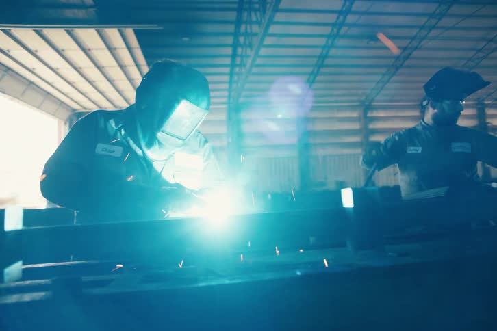 Slow motion shot of two welders in a dark industrial warehouse working on long metal beams, creating bright flashes and flying sparks