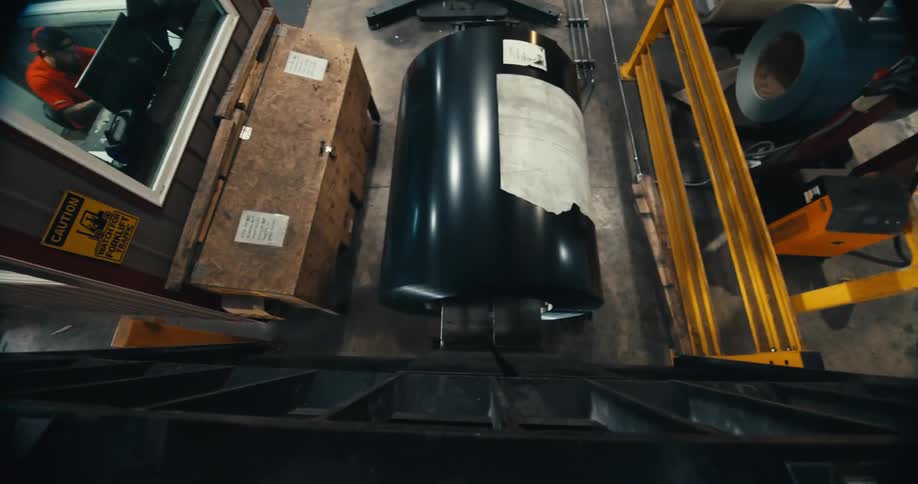 High-angle point of view shot of a forklift lifting a large, heavy coil of black sheet metal in an industrial warehouse