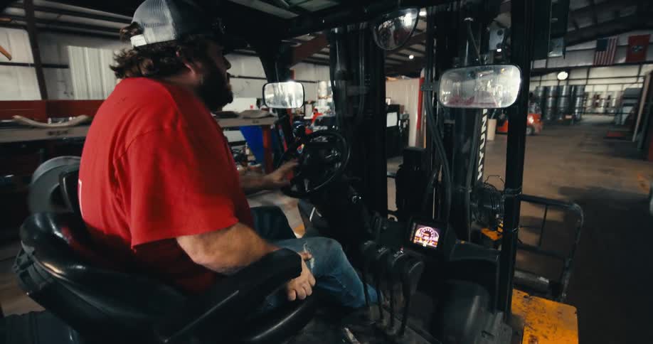 A point-of-view shot from inside a forklift as a bearded man in a red shirt drives through an industrial warehouse