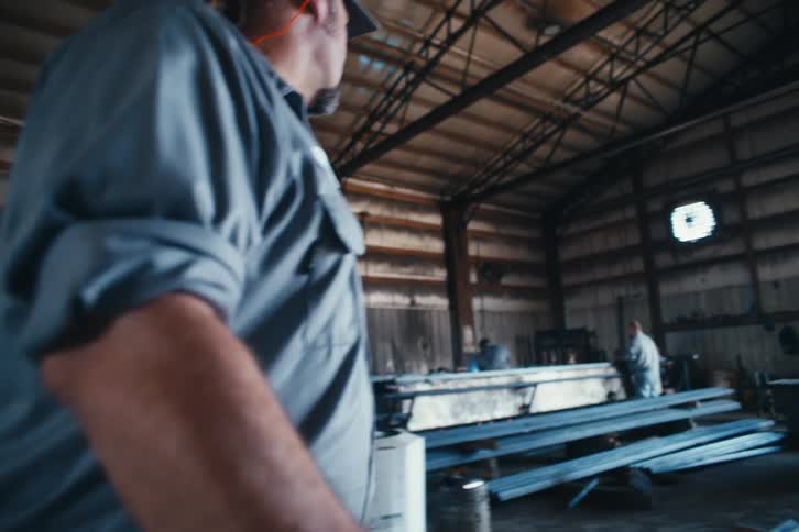 A blue-collar worker named Adam walks through a busy industrial warehouse in slow motion, giving a thumbs up to his team