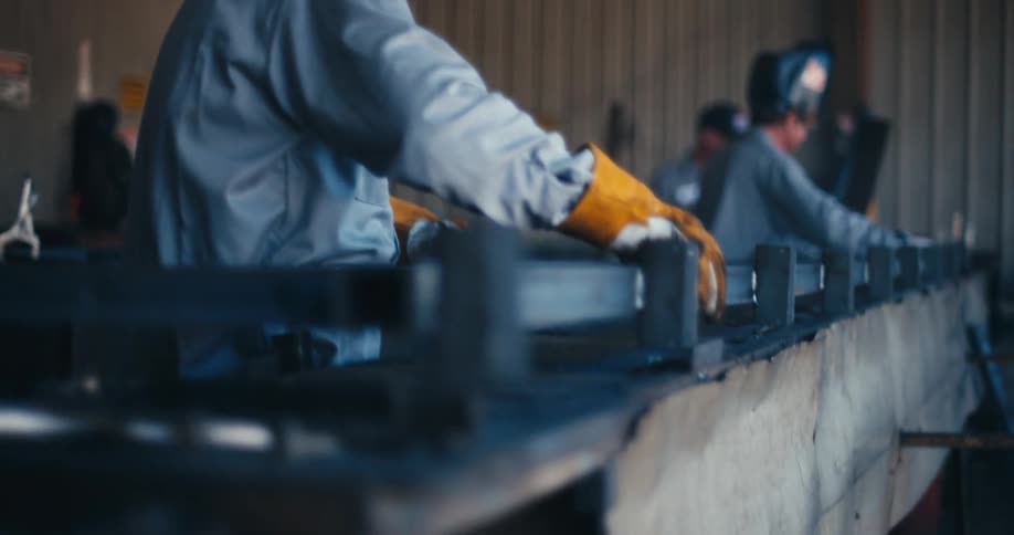 A welder wearing protective gear carefully places a metal railing onto a jig in a busy fabrication workshop