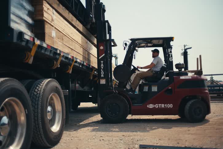 A worker in slow motion operates a forklift to carefully load a stack of heavy metal trusses onto a flatbed truck at an industrial yard