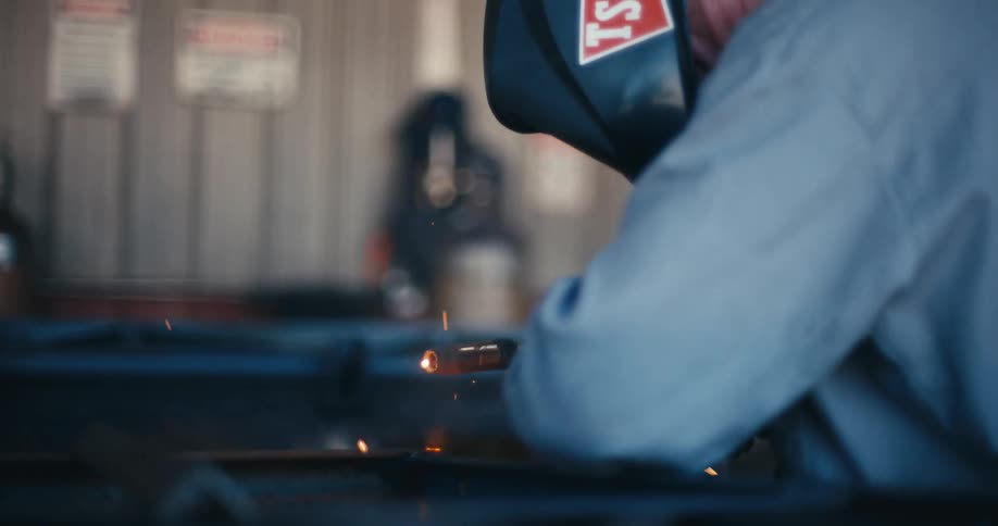 A professional welder wearing a protective helmet carefully fuses metal, generating a cascade of bright sparks and intense blue light in a slow-motion close-up