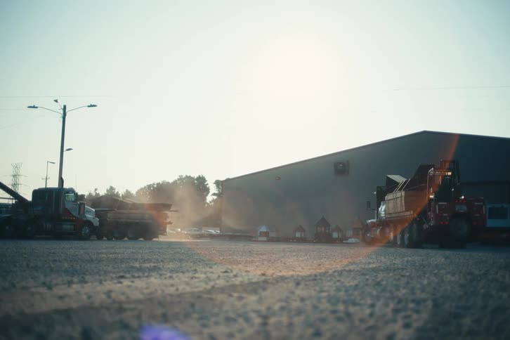 A large semi-truck backs into an industrial lot kicking up dust as the sun shines brightly in the background