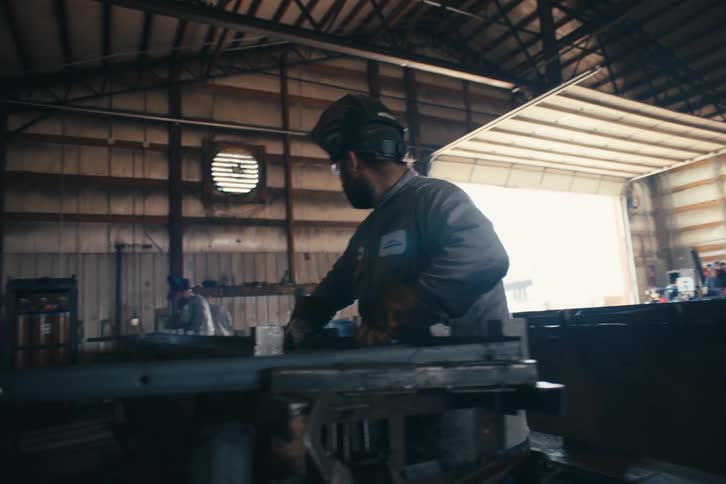 A focused welder meticulously assembles and welds a large metal truss inside an industrial workshop