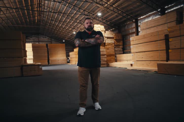 A confident, tattooed worker with his arms crossed looks at the camera while standing in a large lumber warehouse