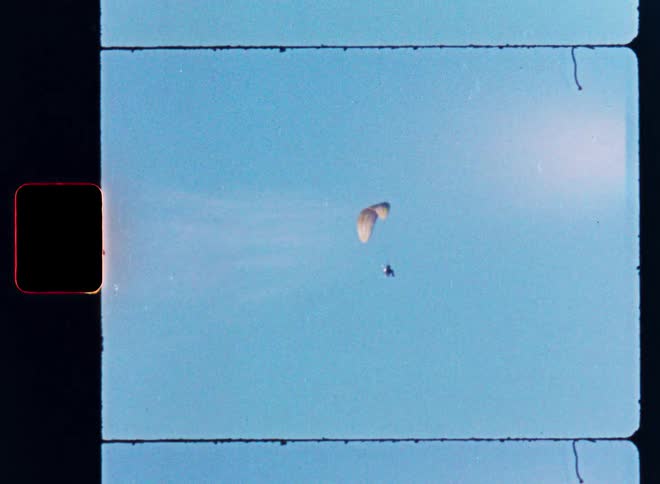 A vintage 8mm film scan shows a single paraglider soaring peacefully in a clear blue sky