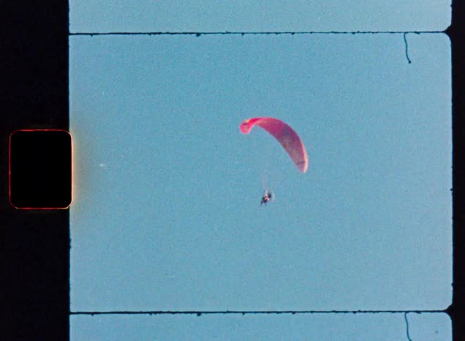 Vintage 8mm film footage shows a lone paraglider drifting across a clear blue sky