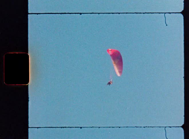 A paraglider with a red chute soars through a clear blue sky in this vintage Super 8 film footage
