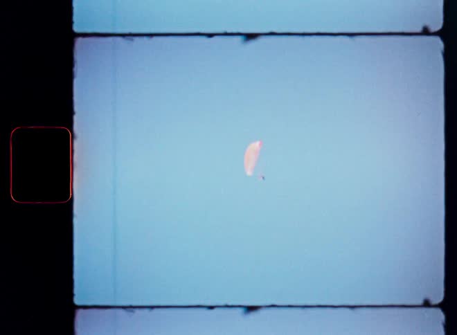A vintage 8mm film captures a lone paraglider soaring peacefully in the middle of a vast, clear blue sky