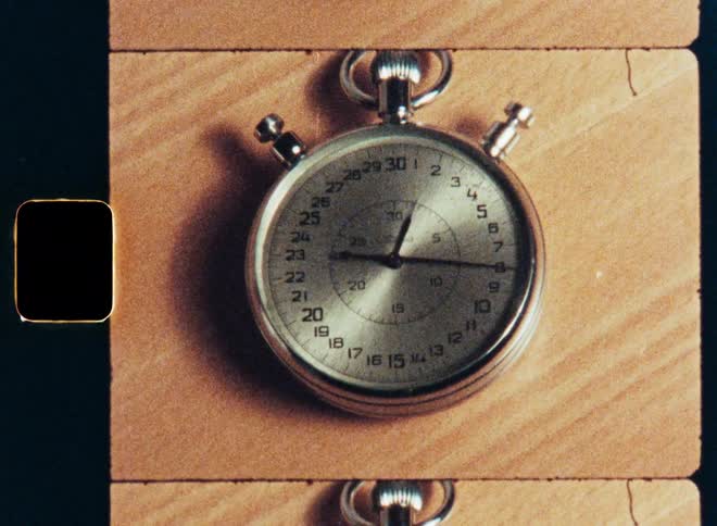 A vintage film scan slowly zooms in on the face of an old, classic stopwatch on a wooden surface