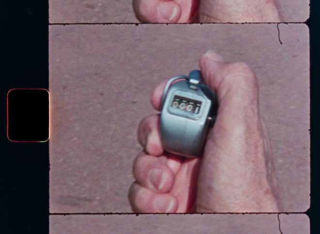 An older person's hand holds a vintage mechanical tally counter, clicking it once from zero to one in a grainy film close-up