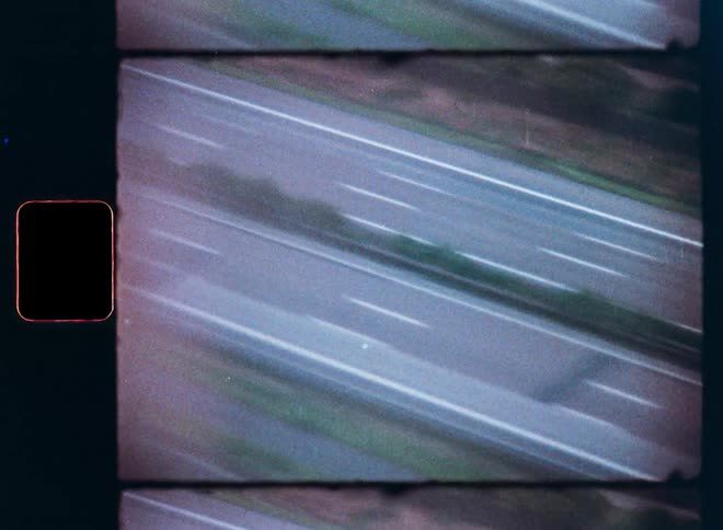 Grainy vintage film captures a high-angle, blurry view of traffic speeding down a highway, with sprocket holes visible