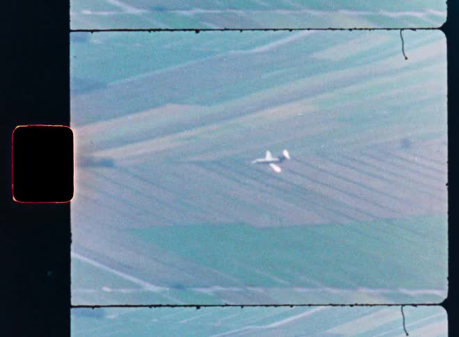 Vintage 8mm film shows an aerial view of a military jet firing on a target over rural fields