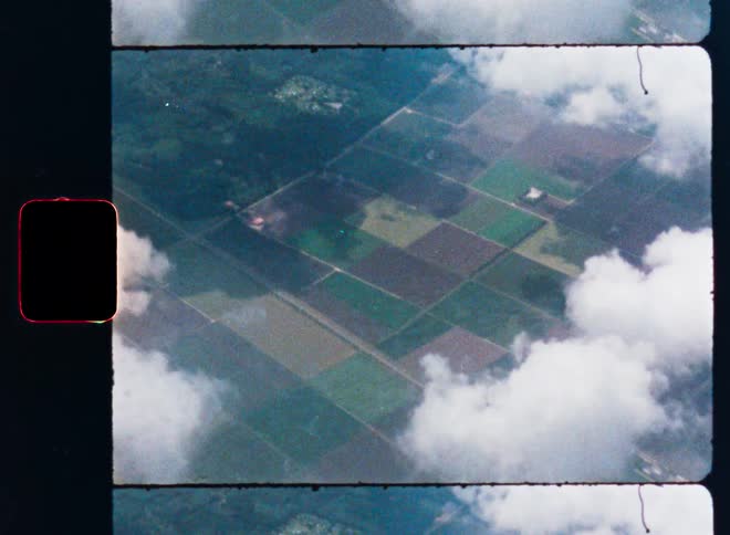A vintage 8mm film scan shows an aerial view of farmlands and forests below fluffy white clouds