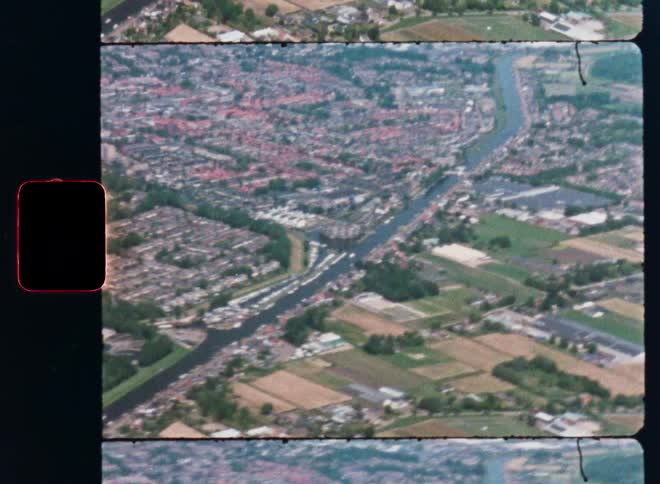 Vintage 8mm film shows a high-angle aerial view of a European city divided by a canal and surrounded by green fields