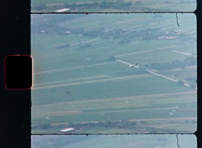 A vintage 8mm film captures an aerial view of a glider soaring peacefully over a patchwork of green farm fields