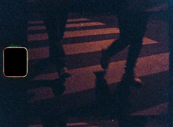 A grainy, vintage 8mm film captures the silhouetted legs and shadows of pedestrians crossing a street at night in a double exposure