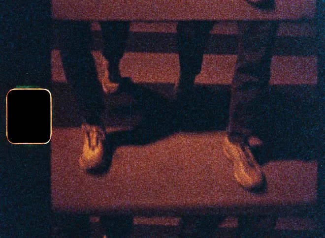 A grainy, low-angle vintage film shot of people's feet in sneakers walking across a crosswalk at night