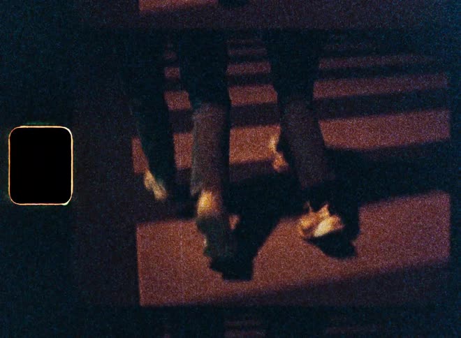 A grainy, vintage-style shot shows the legs and feet of people walking across a striped crosswalk at night, with a film burn at the end