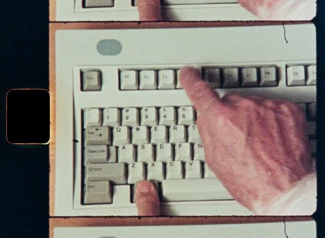 An overhead view on grainy vintage film shows a person hunt-and-peck typing on an old computer keyboard